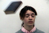 A figure with long brown hair wearing a gray sweater over a light pink and white button-down shirt closes their eyes as a thrown book flies towards their face.
