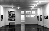 A black-and-white photograph of a room with photographs arranged in a row on the right-hand wall, and in a large grid on the left wall.
