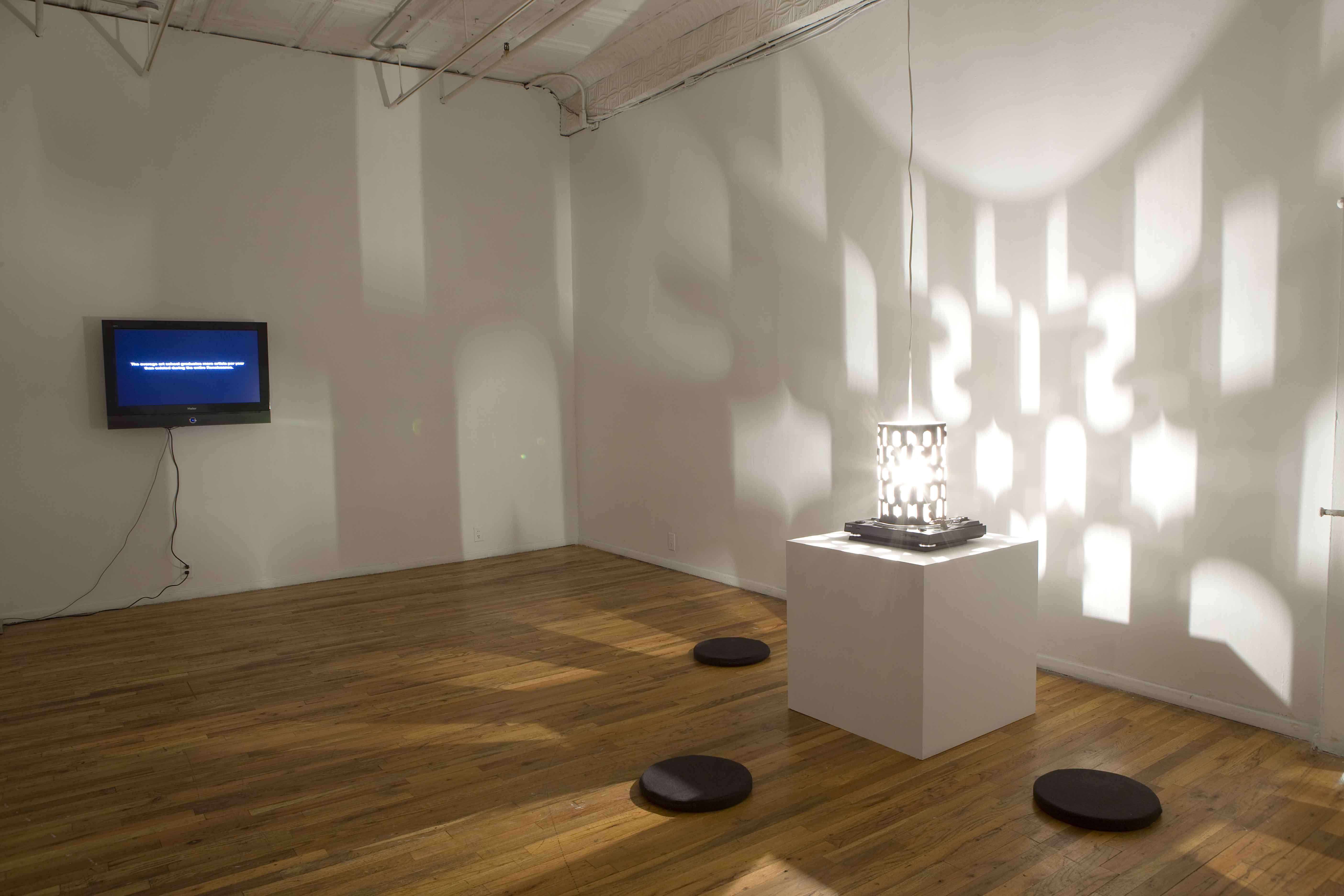 A cylindrical lamp with cutouts in various shapes projects a shadow on white walls. The lamp sits on a white base, surrounded by three circular floor cushions. On the far wall, there is a mounted television screen showing white text.