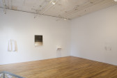 The corner of a room with two white walls. On one wall, there is a white collared shirt on a hanger, a black and white abstract image, and a small white sculpture.