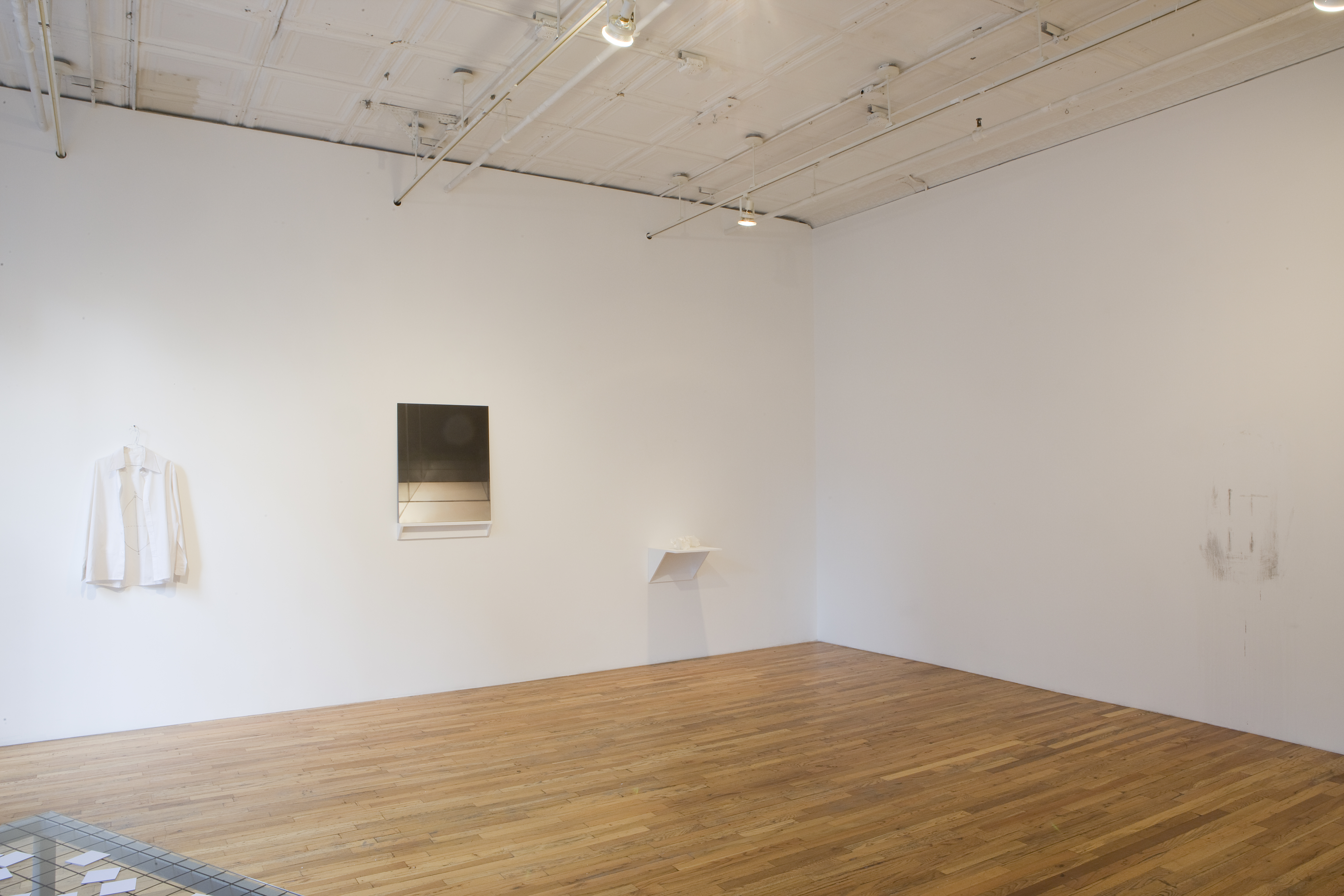 The corner of a room with two white walls. On one wall, there is a white collared shirt on a hanger, a black and white abstract image, and a small white sculpture.