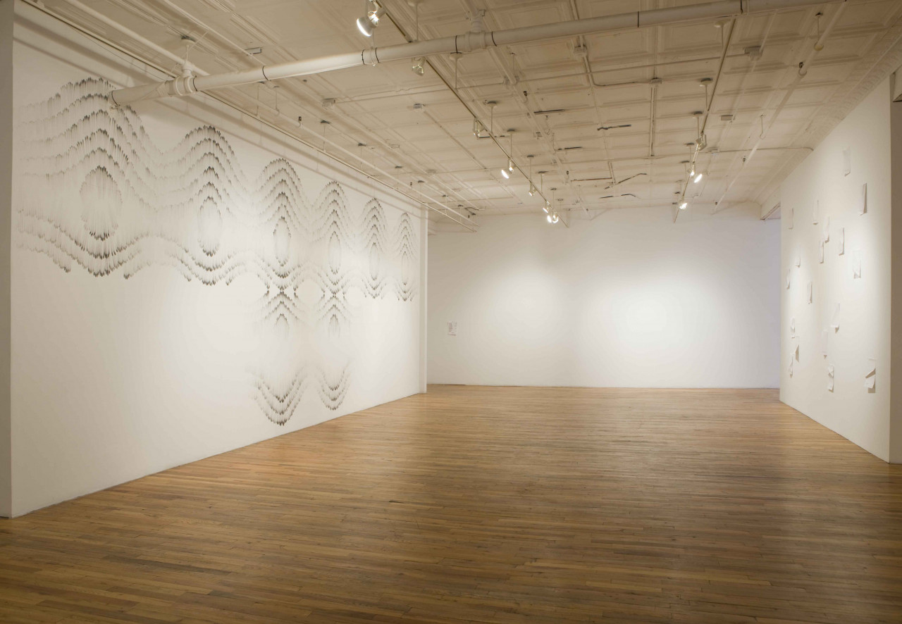 A large, empty room with three white walls. The right wall displays glossy papers folded into geometric shapes. The left wall has symmetric, geometric patterns drawn on it.