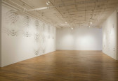 A large, empty room with three white walls. The right wall displays glossy papers folded into geometric shapes. The left wall has symmetric, geometric patterns drawn on it.