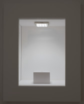 A white plinth in a white room is seen through a rectagular cutout window. There is a small object on the plinth, lit from above by a square, fluorescent light.
