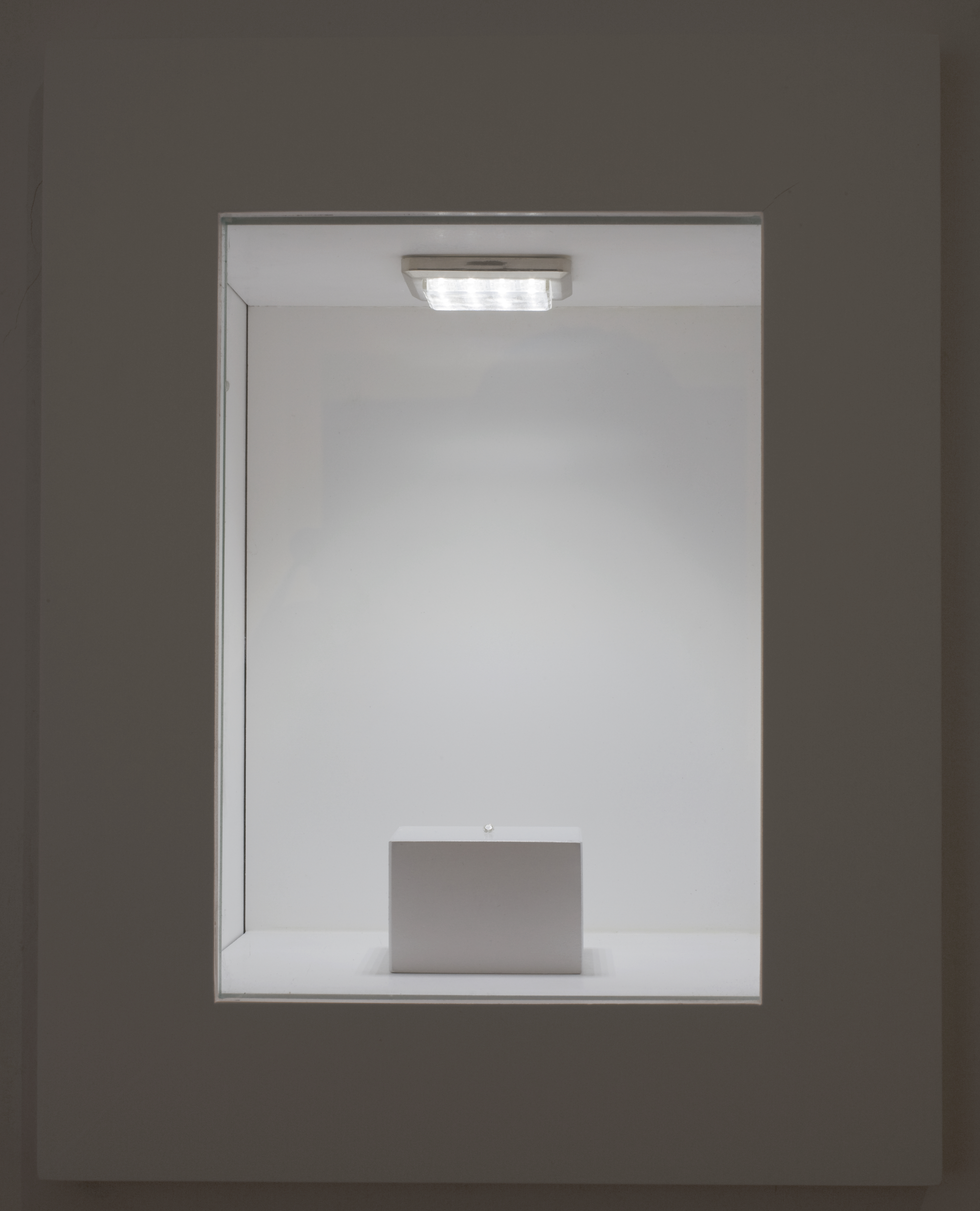 A white plinth in a white room is seen through a rectagular cutout window. There is a small object on the plinth, lit from above by a square, fluorescent light.