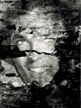 A close-up, black and white image of a face, with parts of the image chipping off and revealing a black backing.