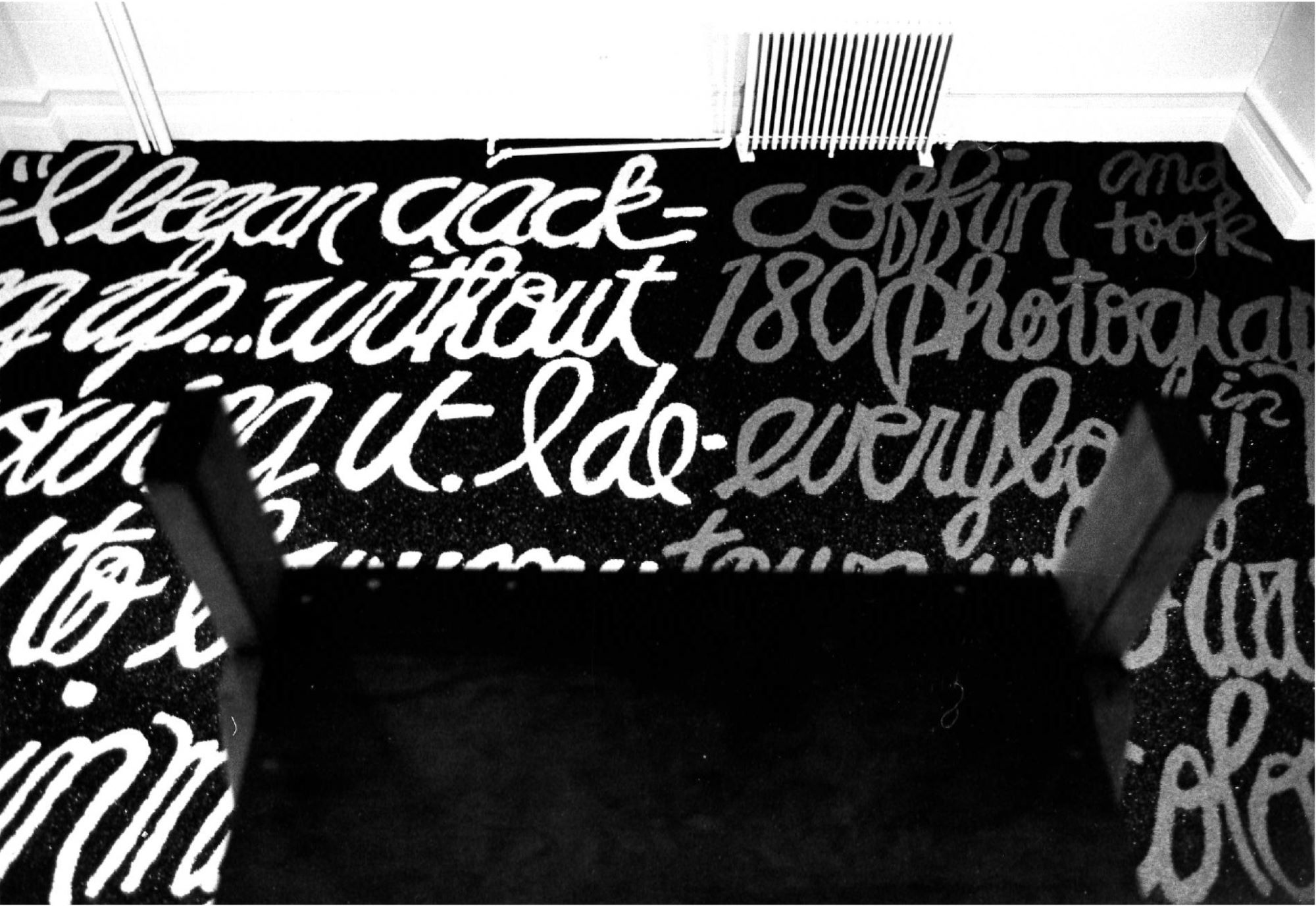 Black and white photograph of cursive white text on a black background on the floor of a room with an upside down chair in the foreground and a heater in the background.