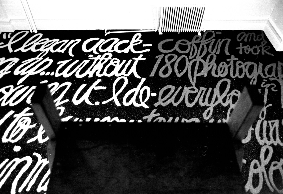 Black and white photograph of cursive white text on a black background on the floor of a room with an upside down chair in the foreground and a heater in the background.