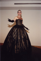 A portrait of a heavily made-up standing person in a ballgown with outstretched arms in black evening gloves. The dark ballgown has rows of light names on it.
