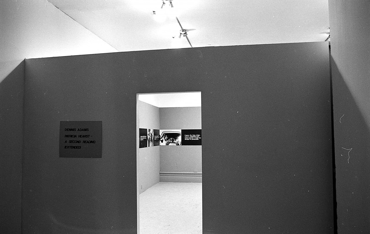 A plaque with text is hung up next to an open doorframe. Through the doorframe, a continuous strip extending around the perimeter of the room is visible. The strip on the walls alternates between photographic stills and black boxes overlaid with white text.