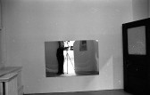 The image of a figure standing next to a camera and tripod is reflected in a mirror hung on a wall.