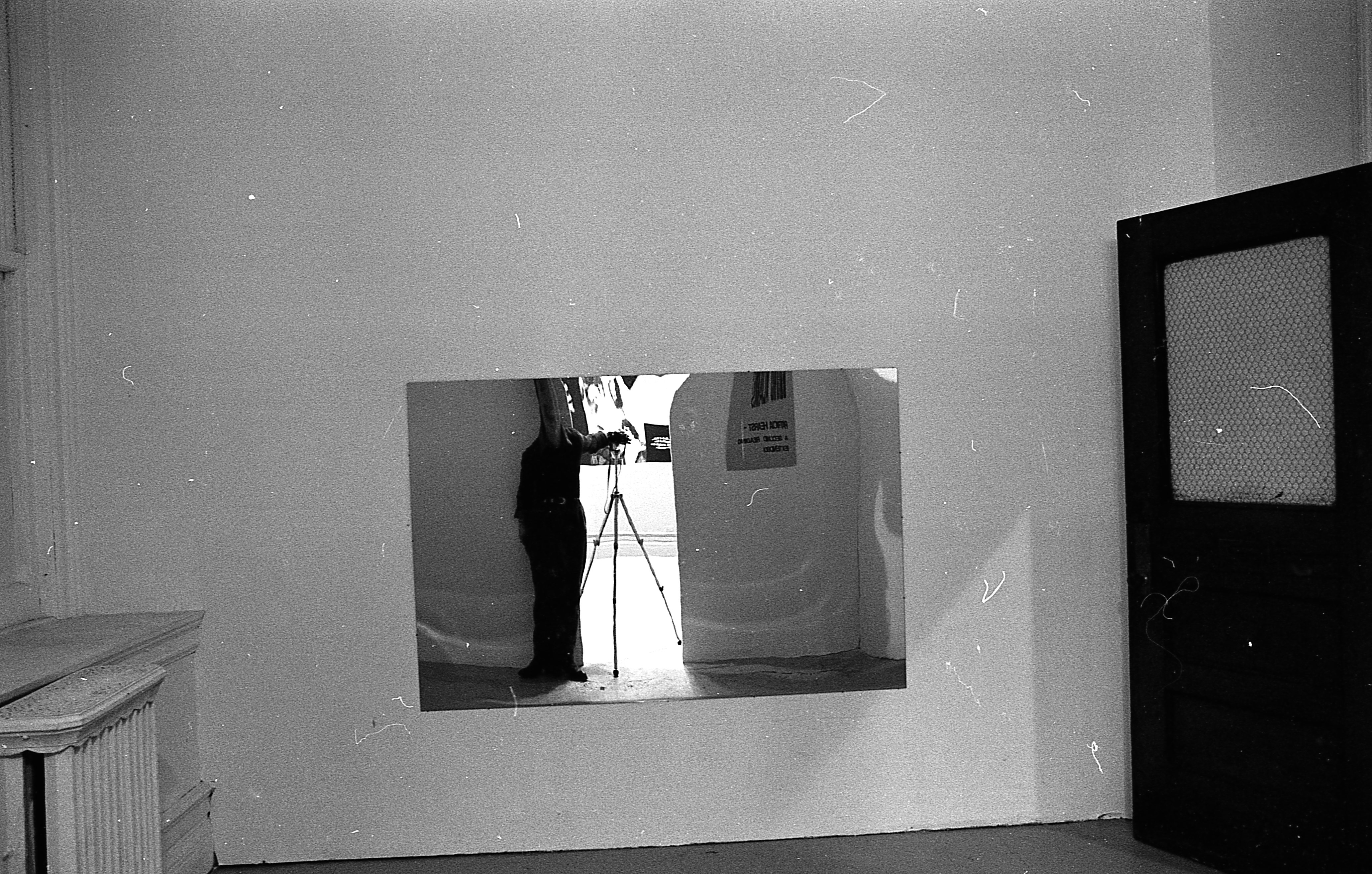The image of a figure standing next to a camera and tripod is reflected in a mirror hung on a wall.