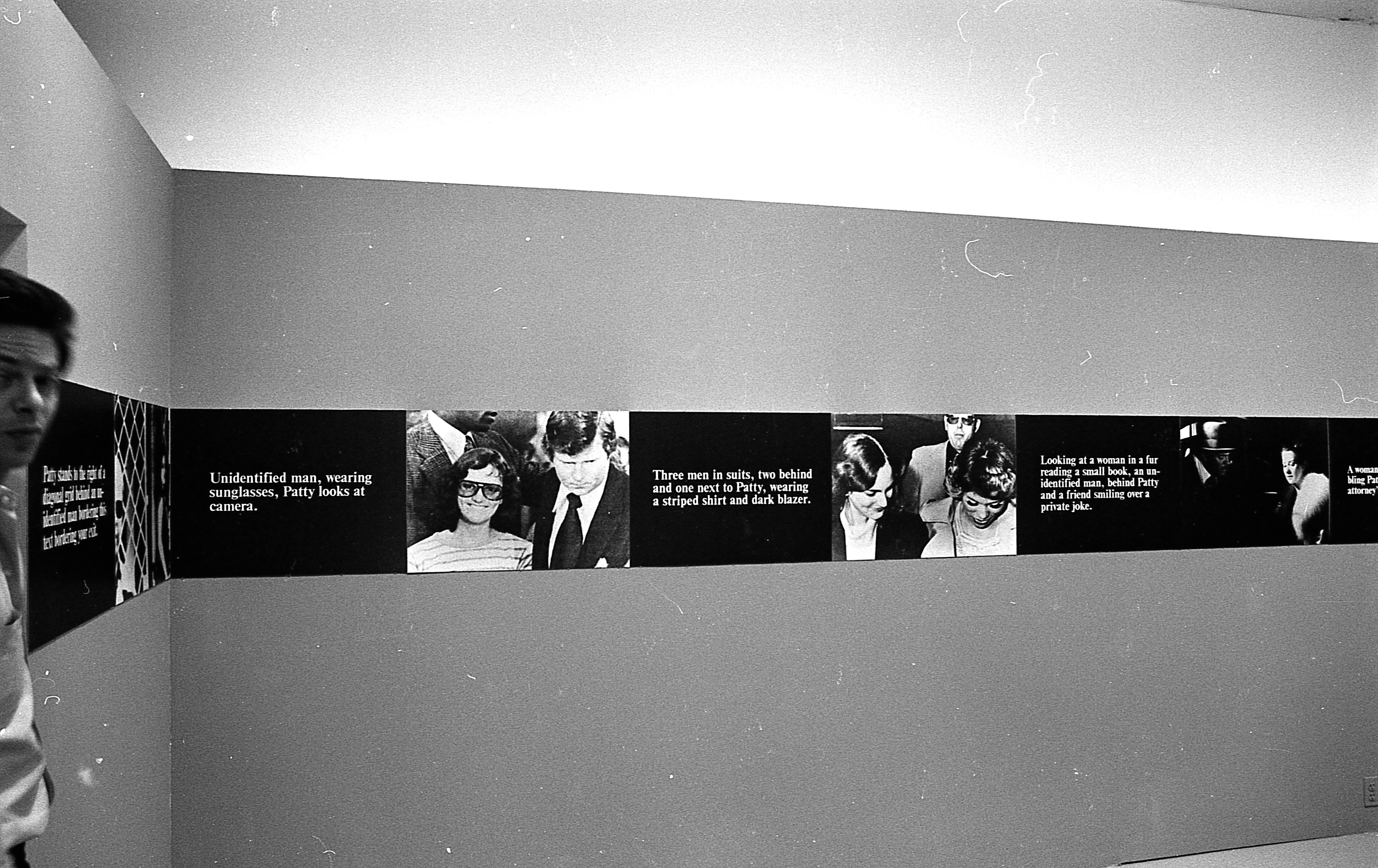 A continuous strip extends around the perimeter of the room, hung up on the walls. The strip alternates between photographic stills and black boxes overlaid with white text.