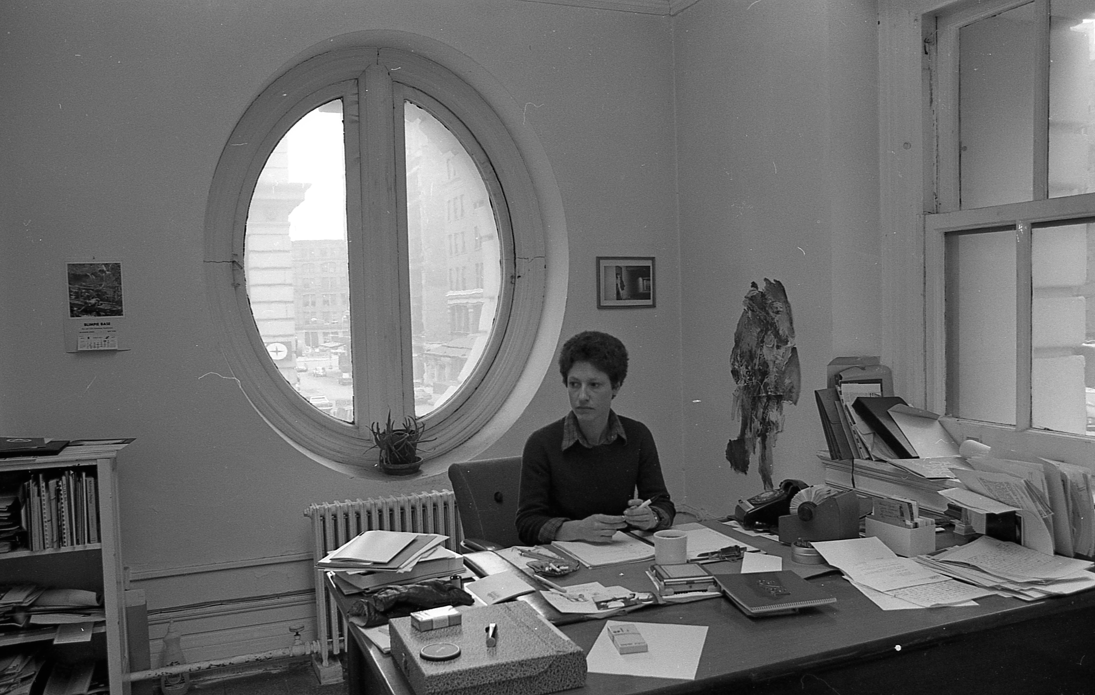 A woman sits at a desk covered with notebooks and documents. She looks off to the side holding a pen by a mug. Behind her, a round window allows light to enter the room.