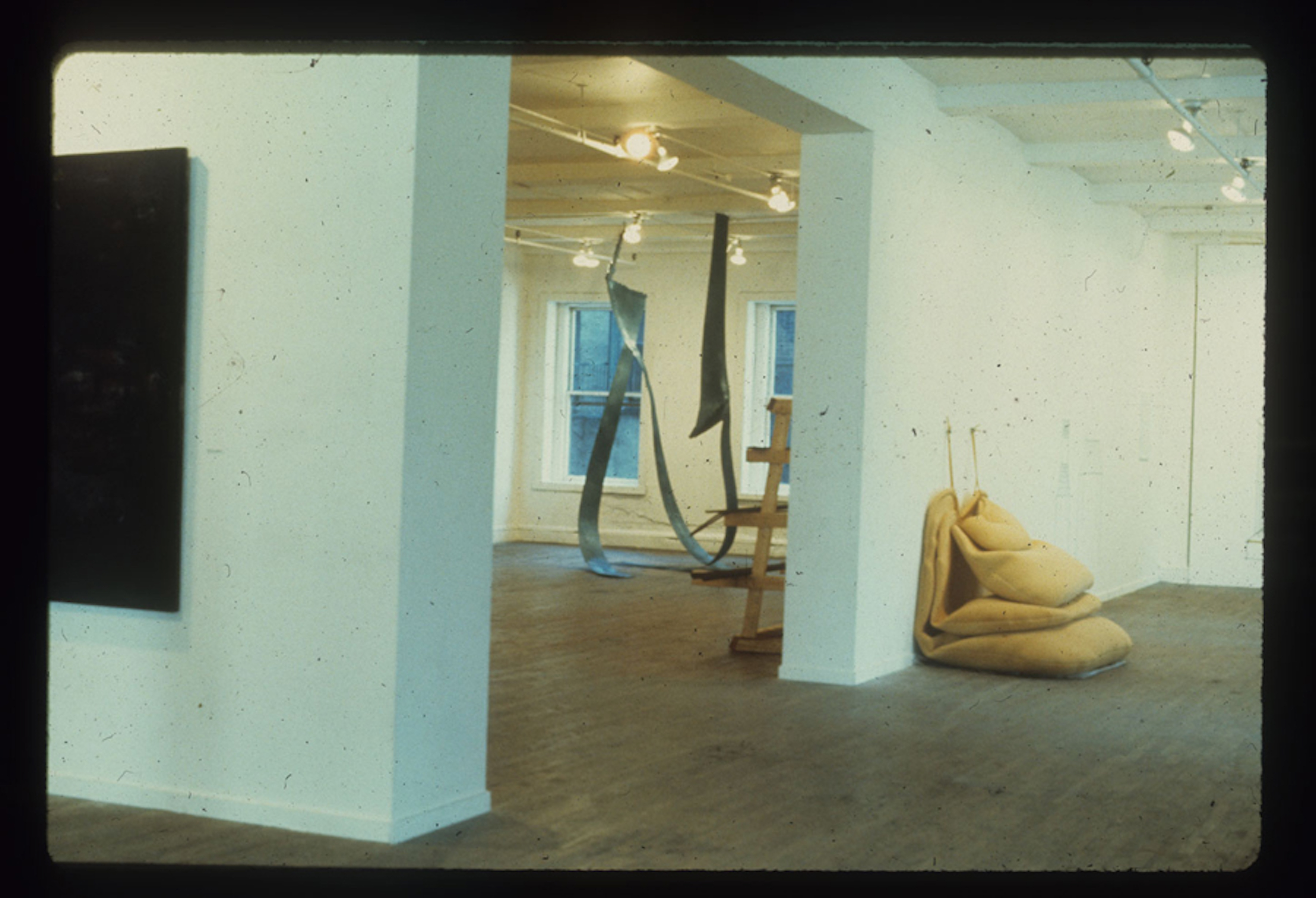 A color installation photograph of a gallery space with a central opening. A black canvas hangs on the wall on the left, and several yellow sculptures are visible on the right.