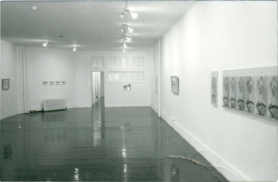 A black-and-white photograph of a gallery space with multiple works, framed and unframed, on the walls.