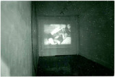 A black-and-white photograph of a gallery space with a video projected on the wall. There appear to be multiple bodies in the video.