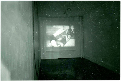 A black-and-white photograph of a gallery space with a video projected on the wall. There appear to be multiple bodies in the video.