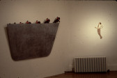 A dimly lit corner of a gallery. A large, gray structure with a slight trapezoidal shape hangs on the corner's left wall. A series of small figurines of men riding on horses emerge in a line along the top edge of the gray form. Hanging on the corner's right wall is a frieze-style figurine of a man in a white shirt and tie that emerges from the wall. He tilts his head back as his arms bend, one extending forward.