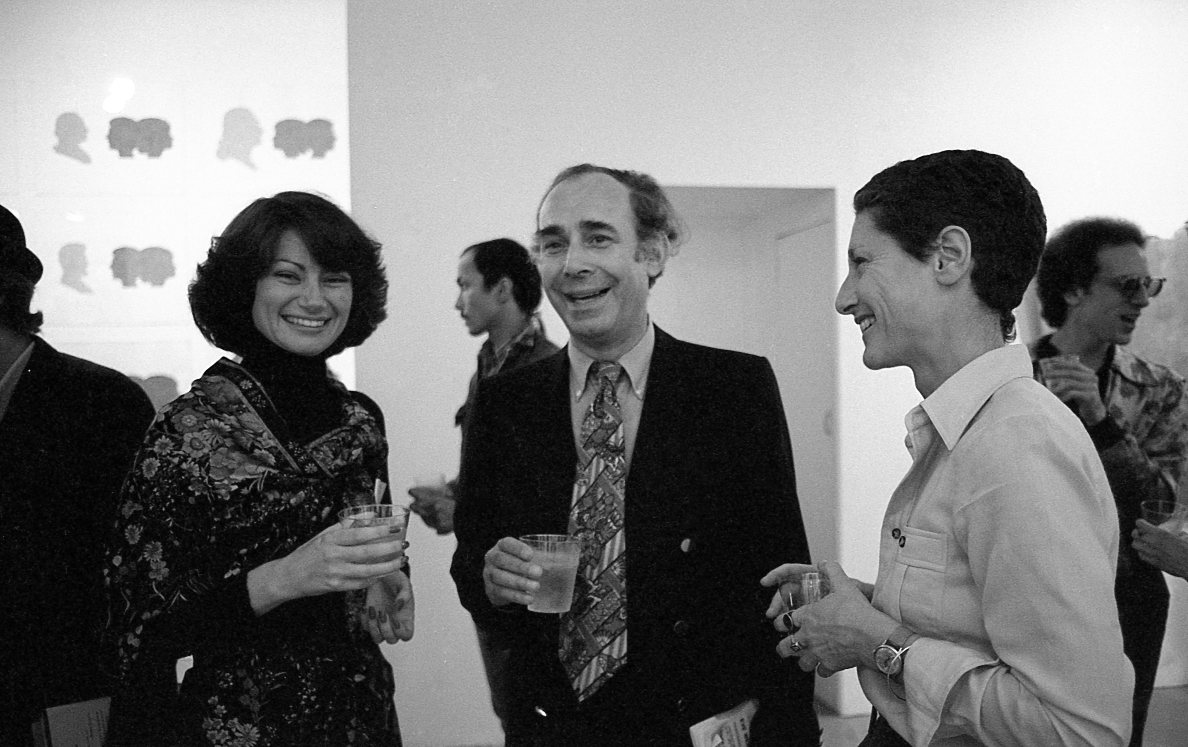 A black and white photograph of three figures standing in a gallery space. They each hold a drink in their hand and are smiling.