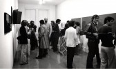 a black and white photograph of a gallery filled with people conversing. The walls are lined with several framed and unframed artworks.