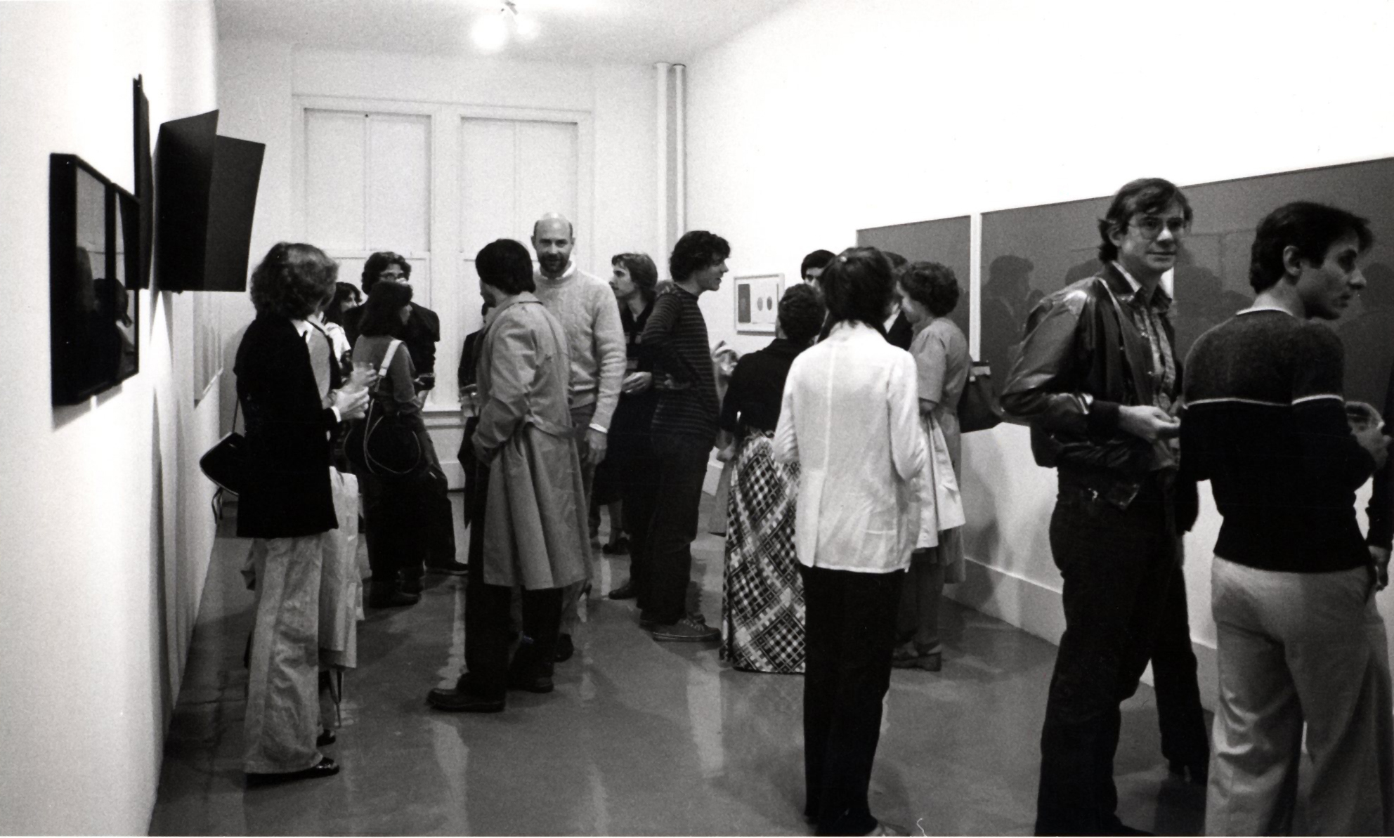 a black and white photograph of a gallery filled with people conversing. The walls are lined with several framed and unframed artworks.