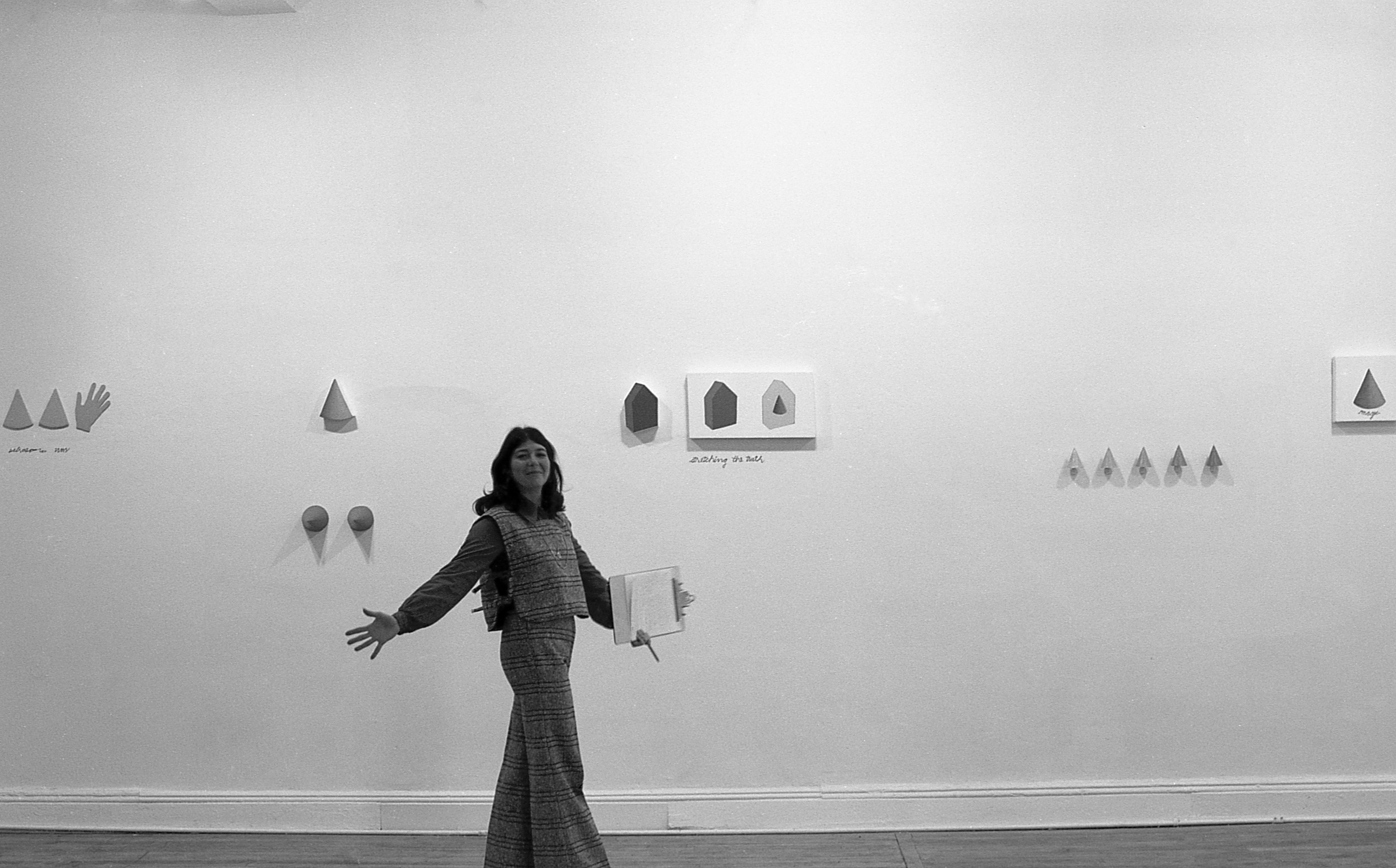 A black and white photograph of a gallery installation. A figure poses in front of a wall with several hanging objects, their arms outstretched to either side.