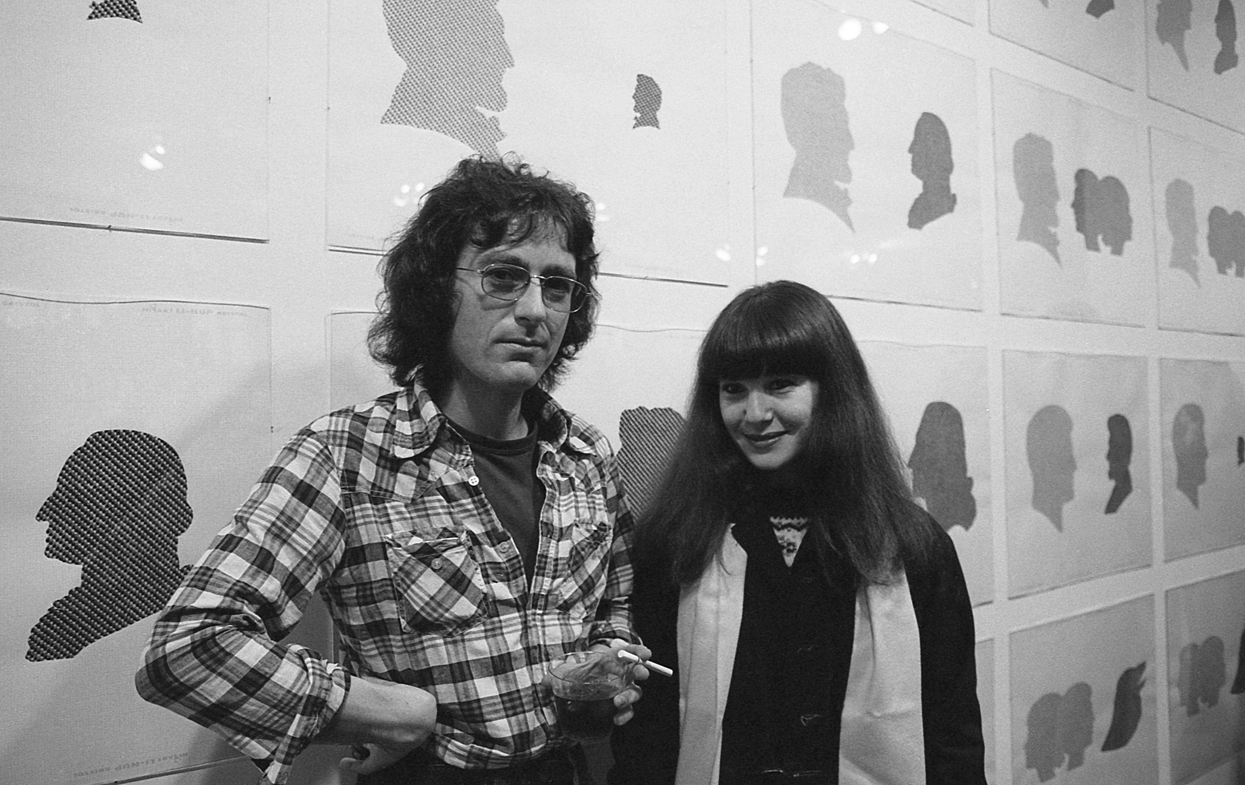 A black and white photograph of two figures standing in front of a wall of framed silhouettes.