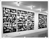 A black and white installation photograph. Four black rectangular panels hang on the wall. Each one is filled with white silhouetted shapes of human figures, and objects.