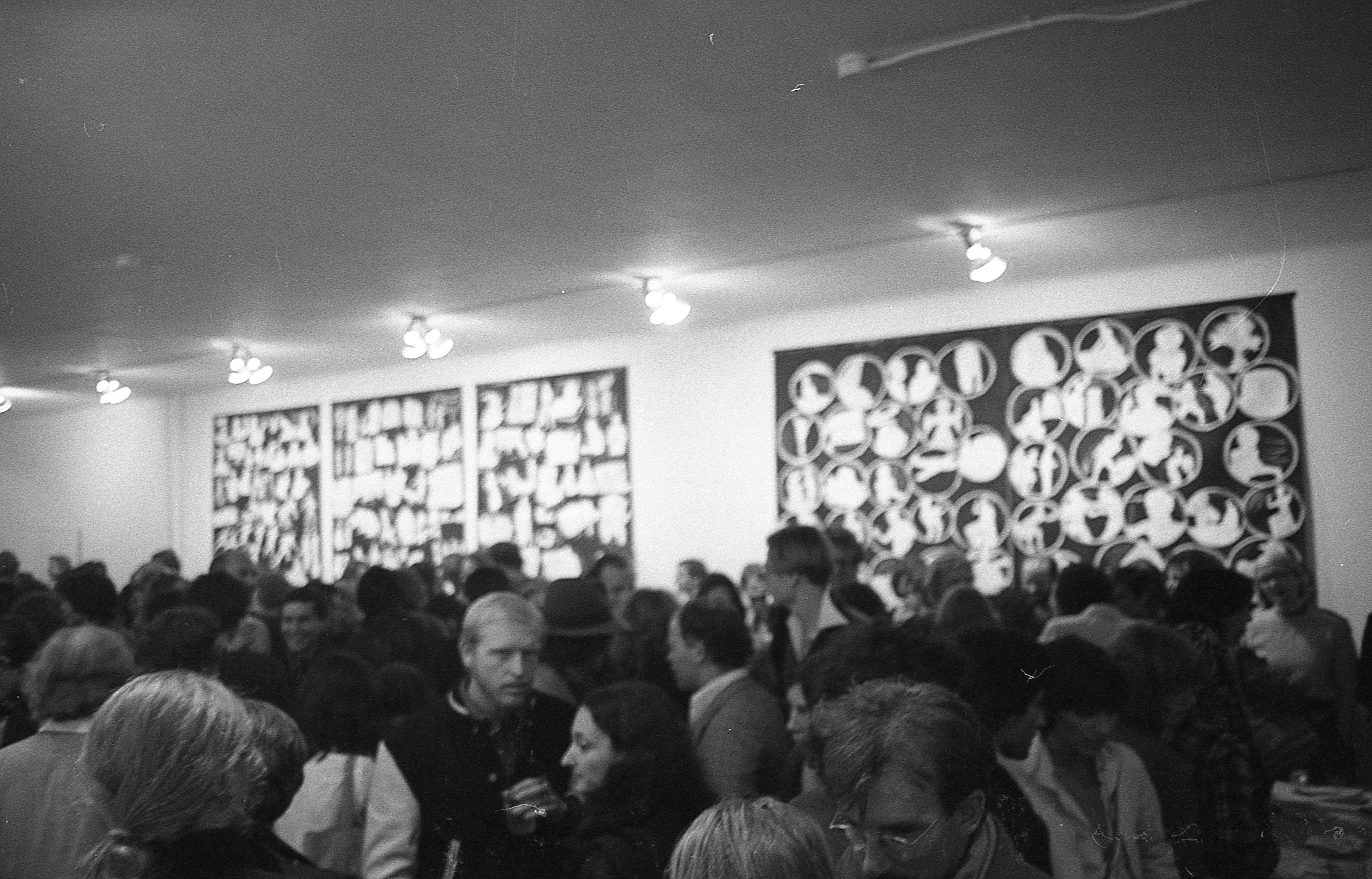 A black and white photograph of a crowded gallery space. In the background, several large black and white wall hangings are visible.