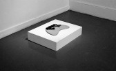 A black and white installation photo of a gallery space. An illuminated low pedastal on the floor holds a crescent shaped object.