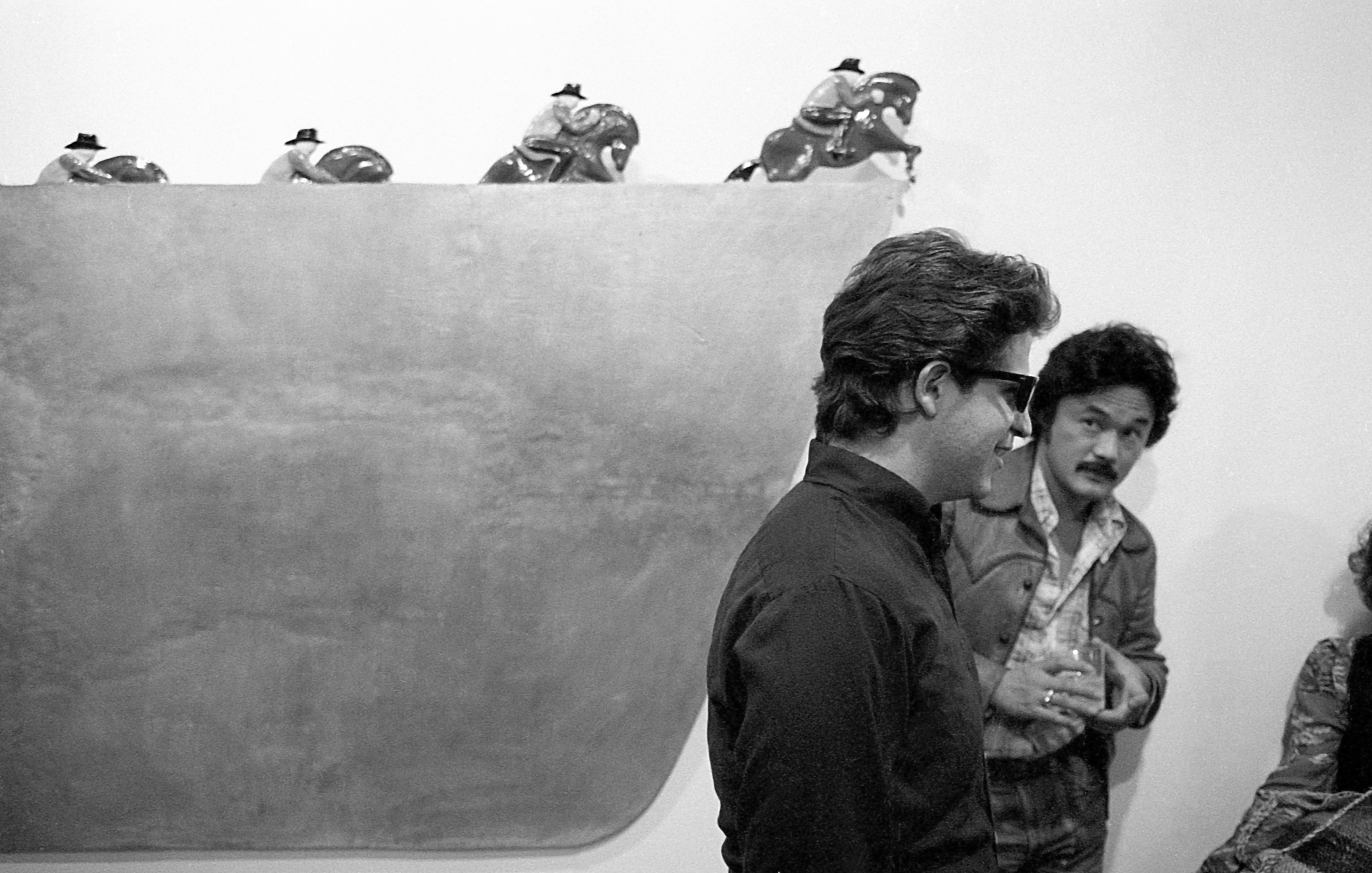 A black and white installation photograph of a gallery space. On the wall, a trapezoidal shape with riders on horseback slowly emerging from the top is visible. On the right, two figures converse.