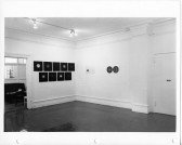 Black and white installation shot of a gallery corner. Attached to the wall are nine square black pieces on the left, and two dark circles on the right.