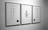 A black and white installation photograph of three framed drawings on a white wall. The center frame is a pared down sketch of a column, and the flanking frames feature feature a simple croshatch of four lines, topped with a fifth.
