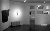 A black and white installation photograph of a gallery space. Several rectangular forms are on the floor, and a large voluminous black rectangle is attached to the wall on the left.