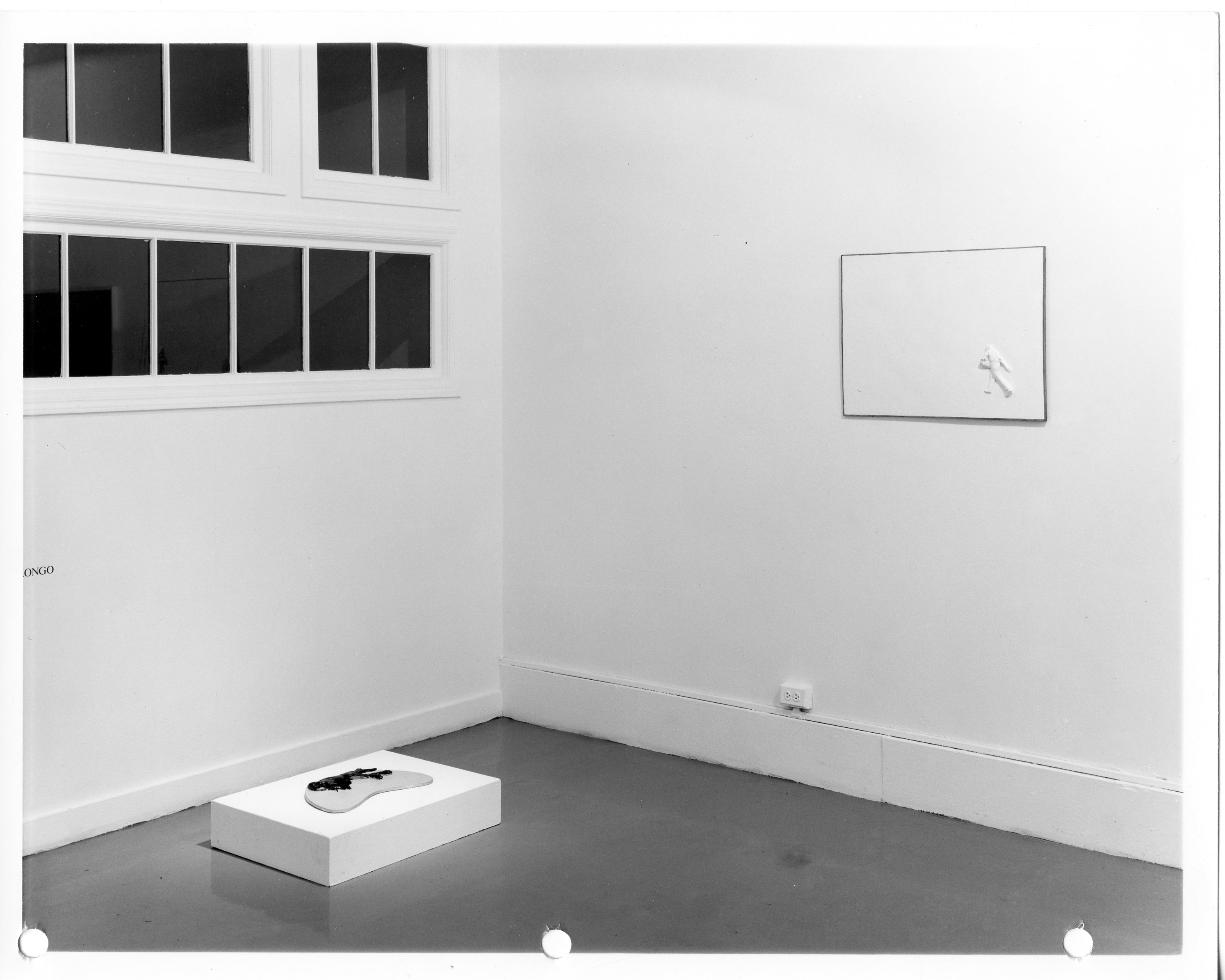 Black and white installation photograph of a gallery corner. On the floor, a crescent shaped object rests on a pedastal. Above, several black panels are attached to the wall.