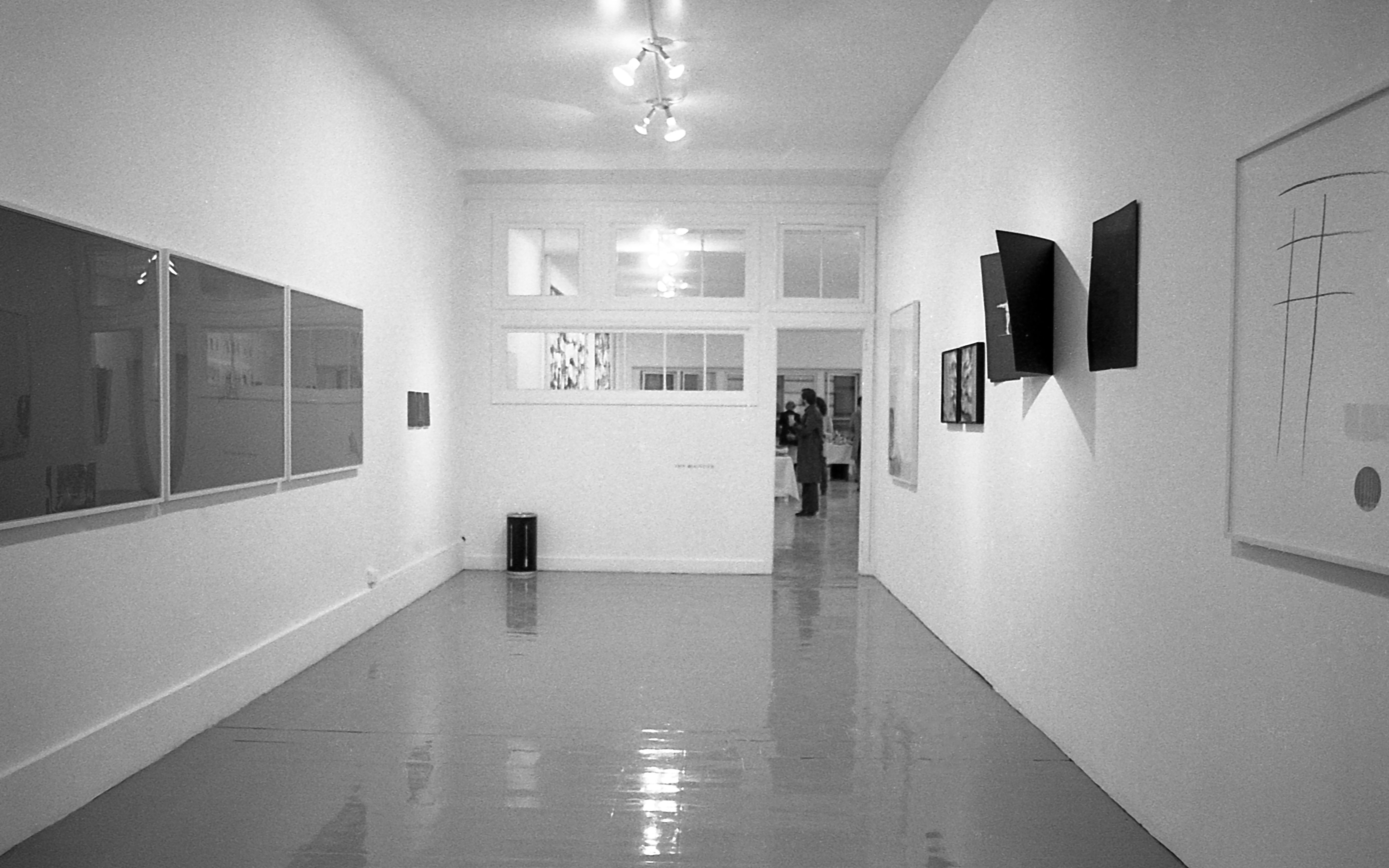 A black and white installation photograph of a gallery space. Several large framed pieces hang on the left and right walls.