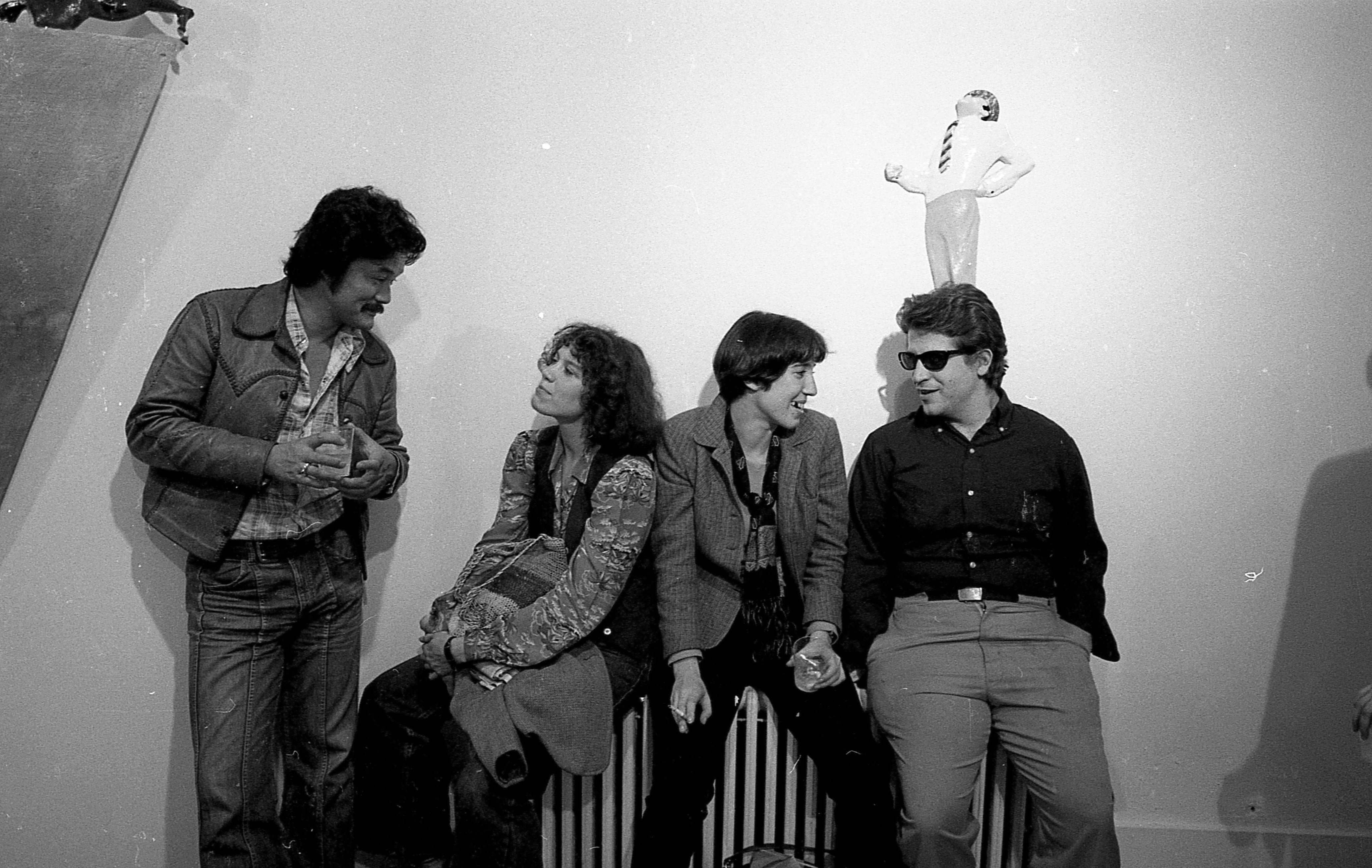 A black and white photograph of an interior with four figures. The left-most figure stands, bending down slightly to speak to another who is seated on a striped bench along with two other people.