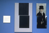 A small square of illegible text, an abstract image of two black squares on grey, and a painting of a person in period dress and a top hat all hang on a blue wall.
