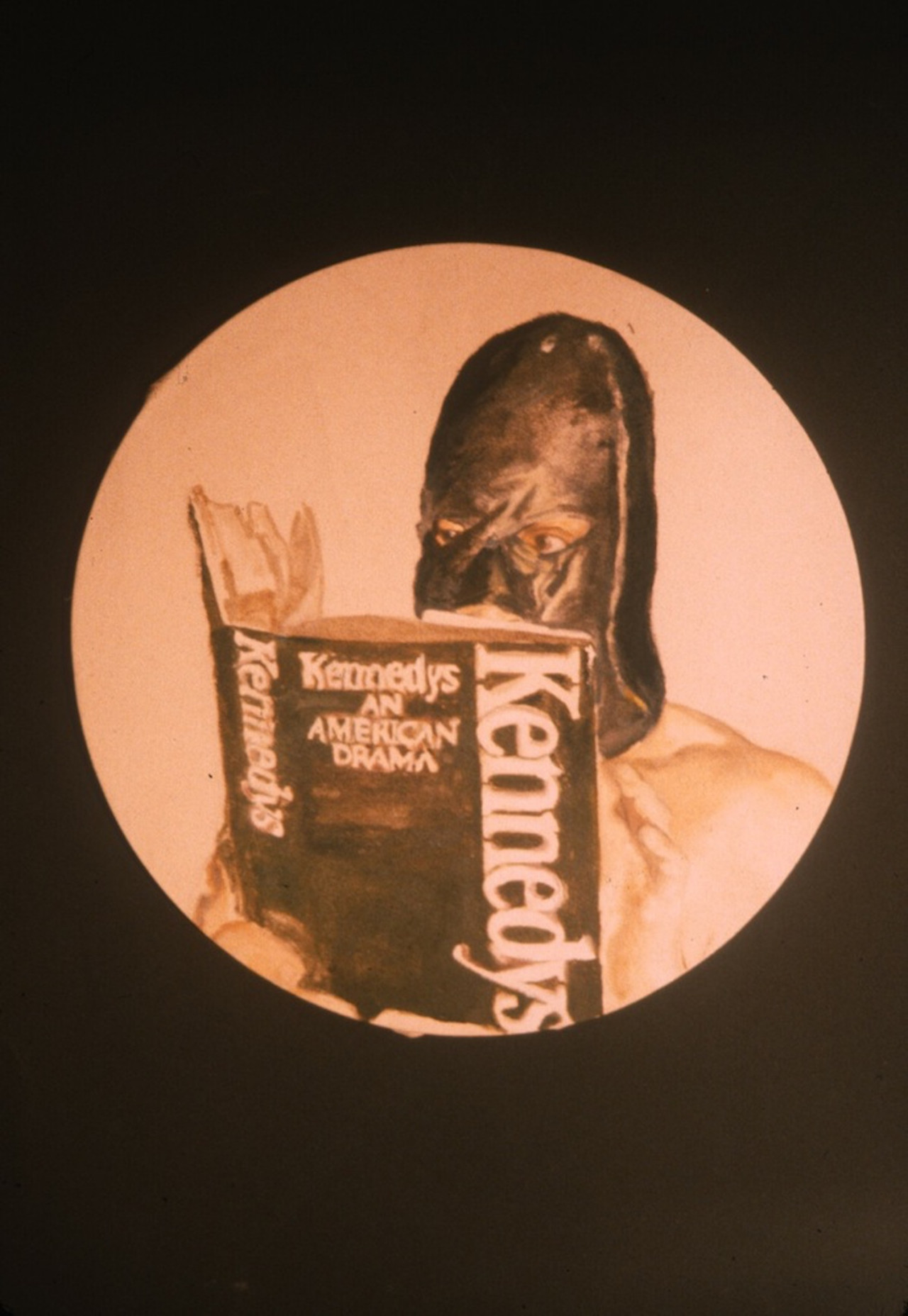 A person wearing a black mask over their head reads a book titled <i>Kennedys An American Dream</i>.