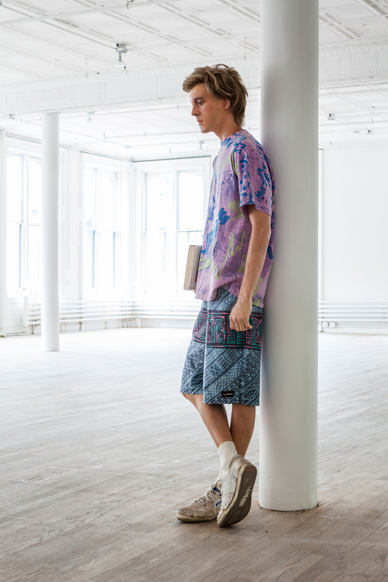 A side view of a sculpture of a man leaning against a white column. He holds a book in his right hand. He is wearing a purple shirt, blue patterned shorts, and white sneakers.