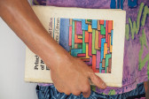A close up view of a sculpture of a man holding a book. The man's hand holds the spine of a book close to his purple shirt.