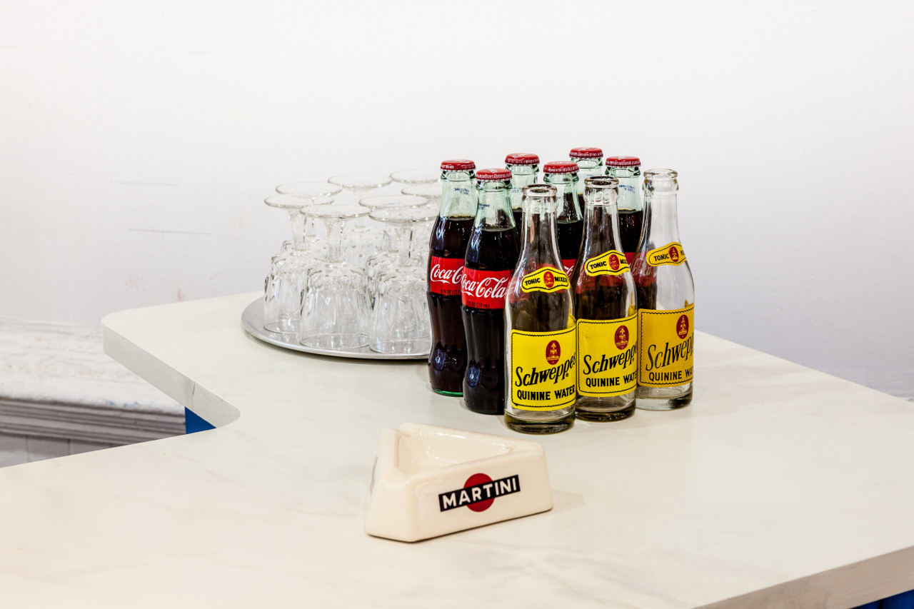 An ashtray with the text, "Martini," three Schweppes bottles, six Coca-Cola bottle, and nine drinking classes arranged face down on a silver platter are displayed on a white counter top.