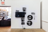 A TV monitor is displayed left of center on a white wall plastered with black-and-white posters that read, 