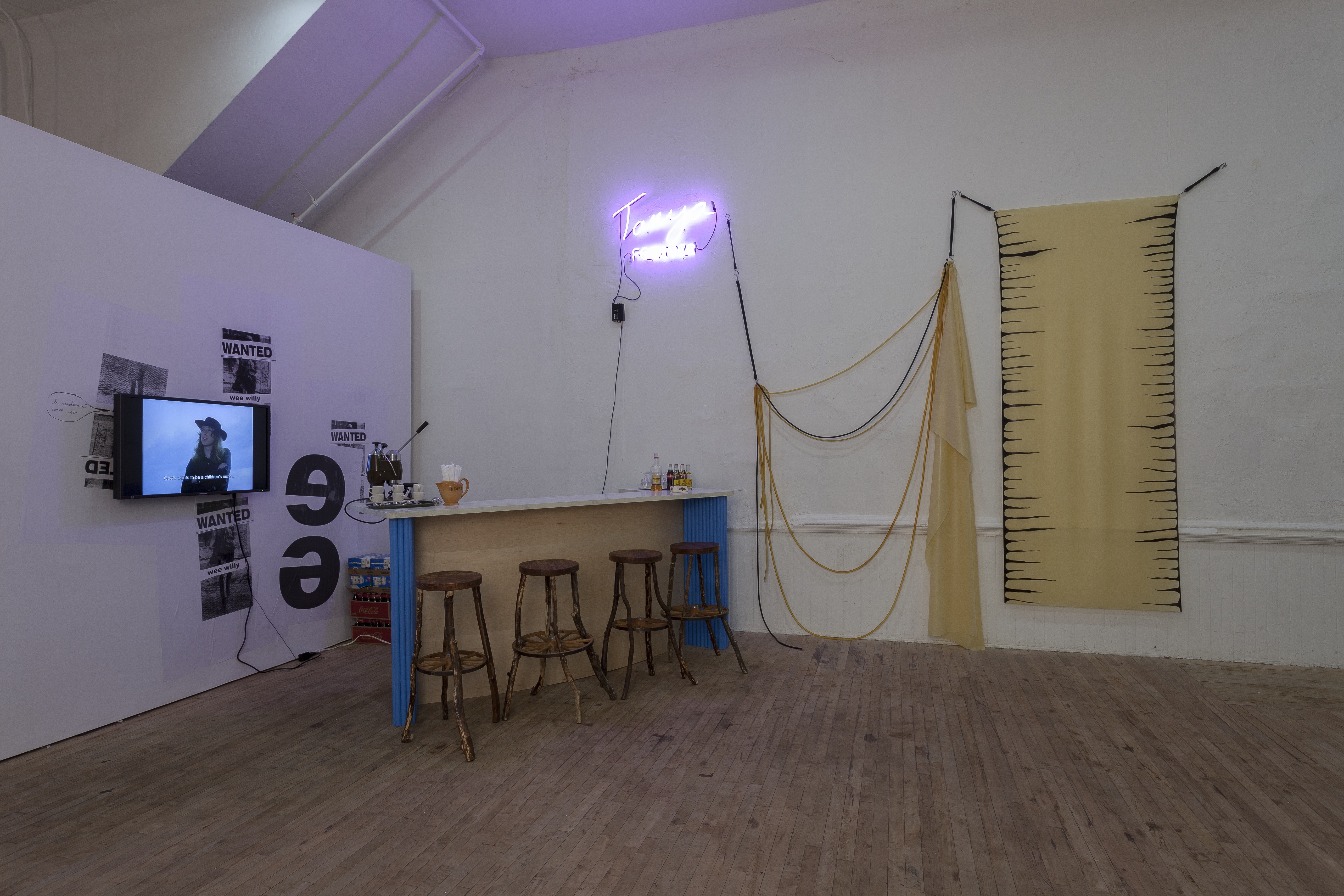A bar with four wooden stools positioned in the corner of a room. A neon sign hangs above to the left of a fiber sculpture. On the left wall, a monitor is displayed on top of plastered posters.