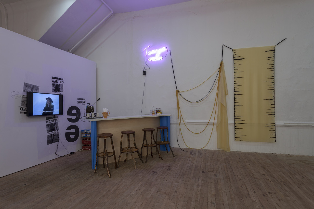 A bar with four wooden stools positioned in the corner of a room. A neon sign hangs above to the left of a fiber sculpture. On the left wall, a monitor is displayed on top of plastered posters.