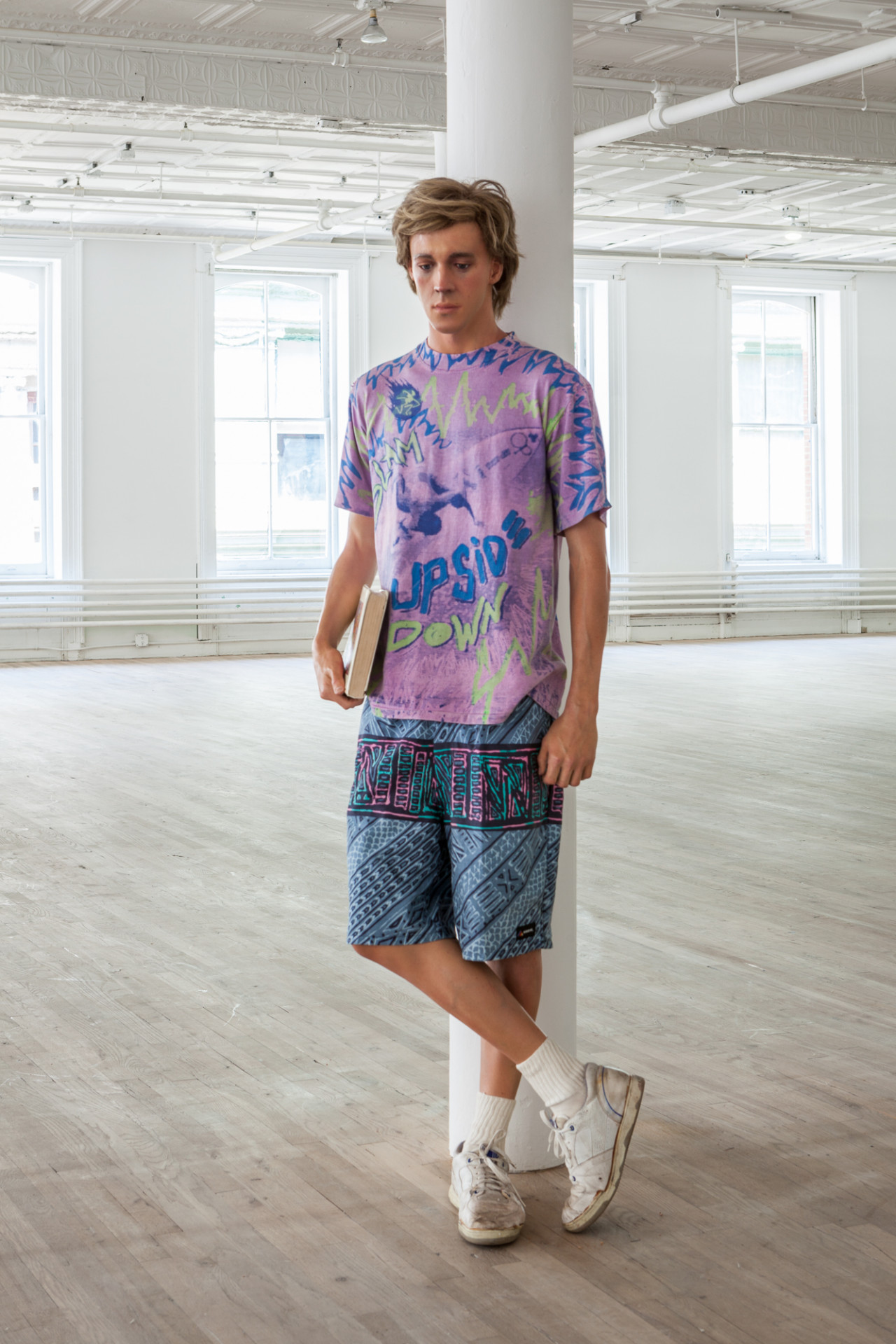 A sculpture of a man holding a book is leaning against a column in an empty, open room. He is wearing a purple shirt, patterned blue shorts, and is crossing his legs, wearing white sneakers.