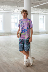 A sculpture of a man holding a book is leaning against a column in an empty, open room. He is wearing a purple shirt, patterned blue shorts, and is crossing his legs, wearing white sneakers.
