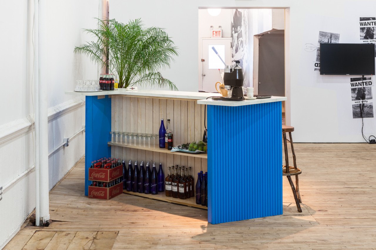 A view from behind an angled bar with blue walls. Shelves full of Coca-Cola, liquor, and glasses are visible. On the counter, more Coca-Cola bottles, glassware, and an esspresso machine. A palm leaf is positioned beyond the counter top.