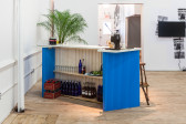 A view from behind an angled bar with blue walls. Shelves full of Coca-Cola, liquor, and glasses are visible. On the counter, more Coca-Cola bottles, glassware, and an esspresso machine. A palm leaf is positioned beyond the counter top.